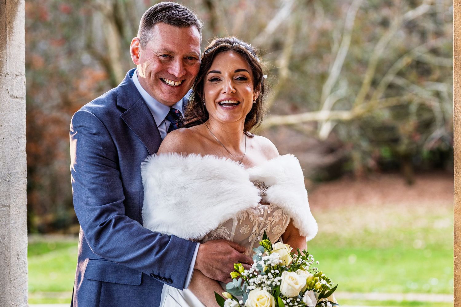 A happy couple embraces outdoors on their wedding day, with the bride wearing a white fur wrap and holding a bouquet.