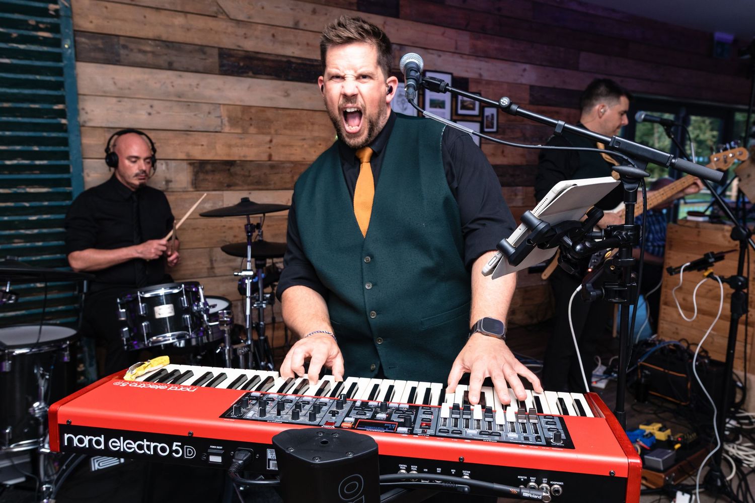 Musician performs energetically on a red Nord keyboard synthesizer in front of a rustic wooden wall.