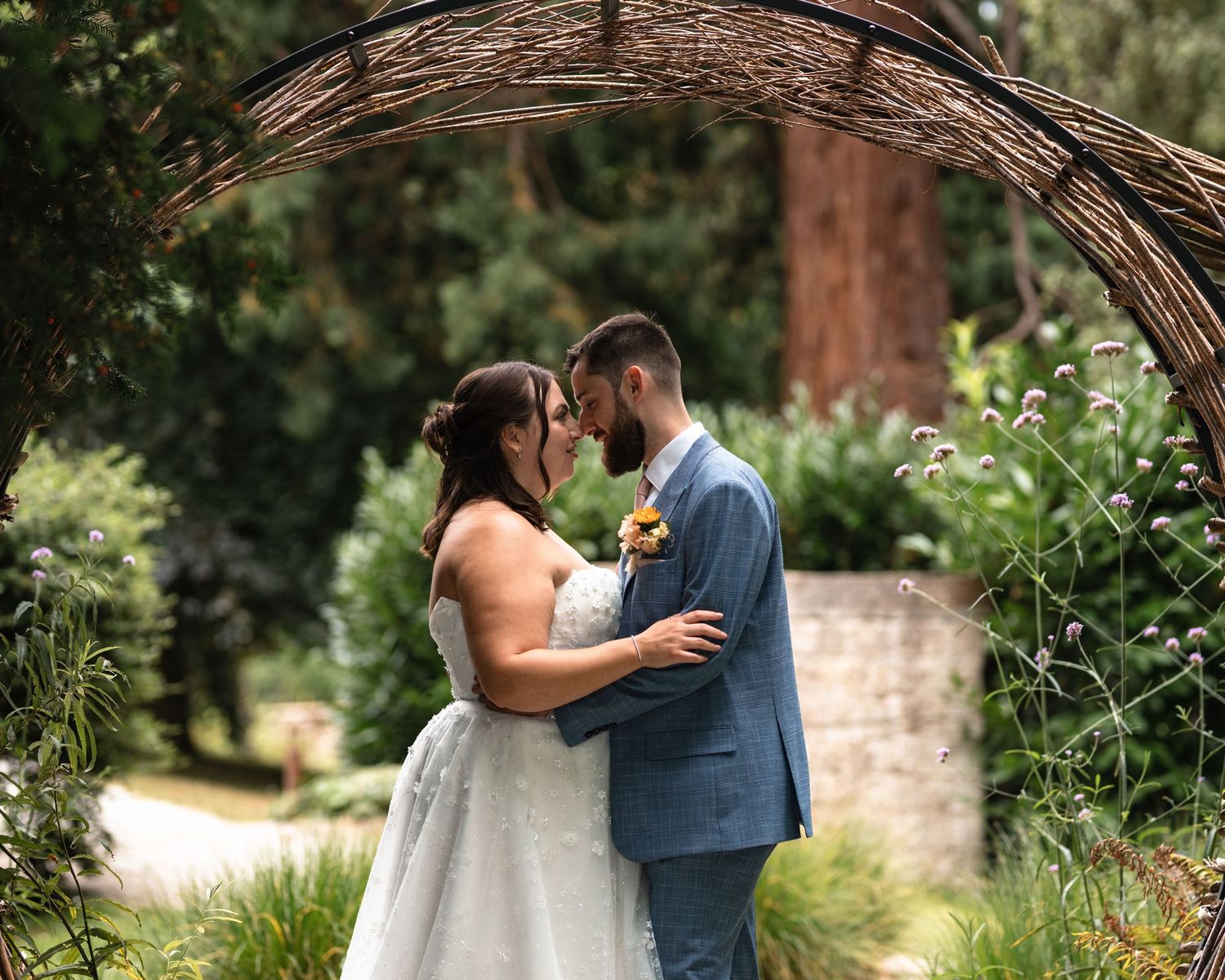 A couple shares an intimate wedding moment under a rustic stone archway in a lush garden setting.