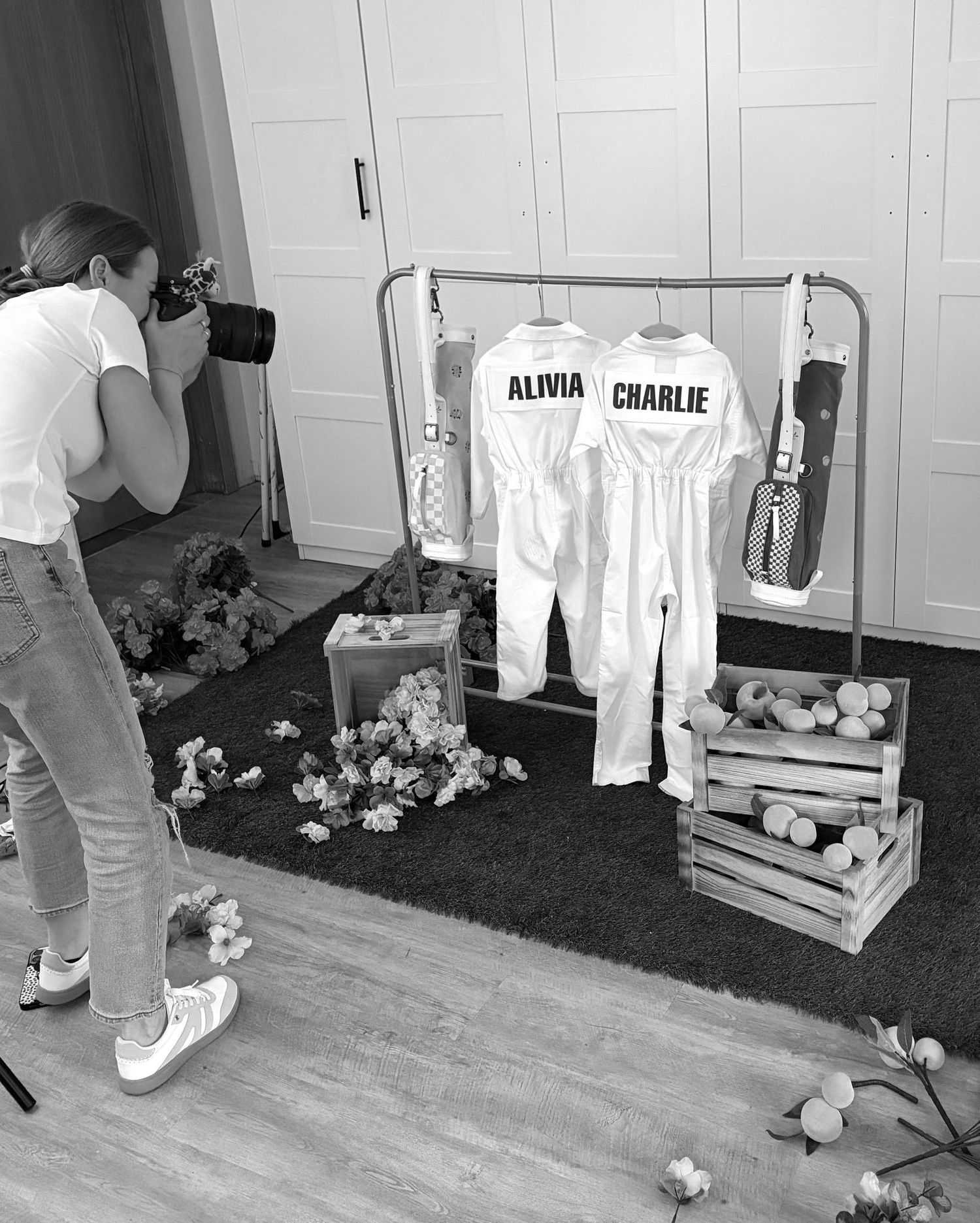 A photographer captures a clothing rack with white outfits labeled 'Alpha Charlie' surrounded by flowers and wooden crates.