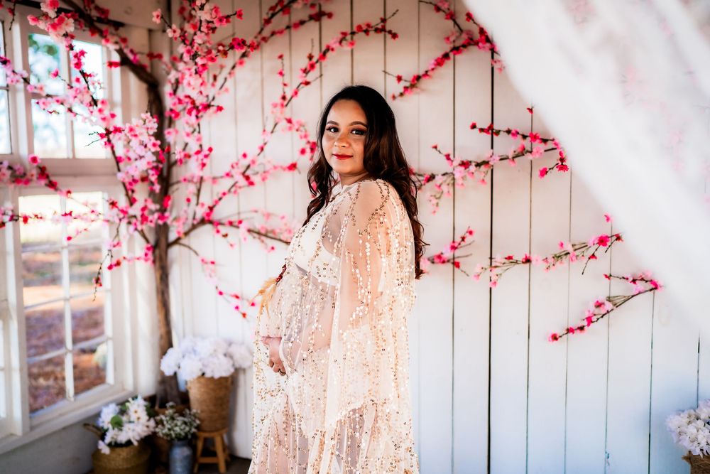 Pregnant woman in lace dress posing before white wall with pink cherry blossom branches.