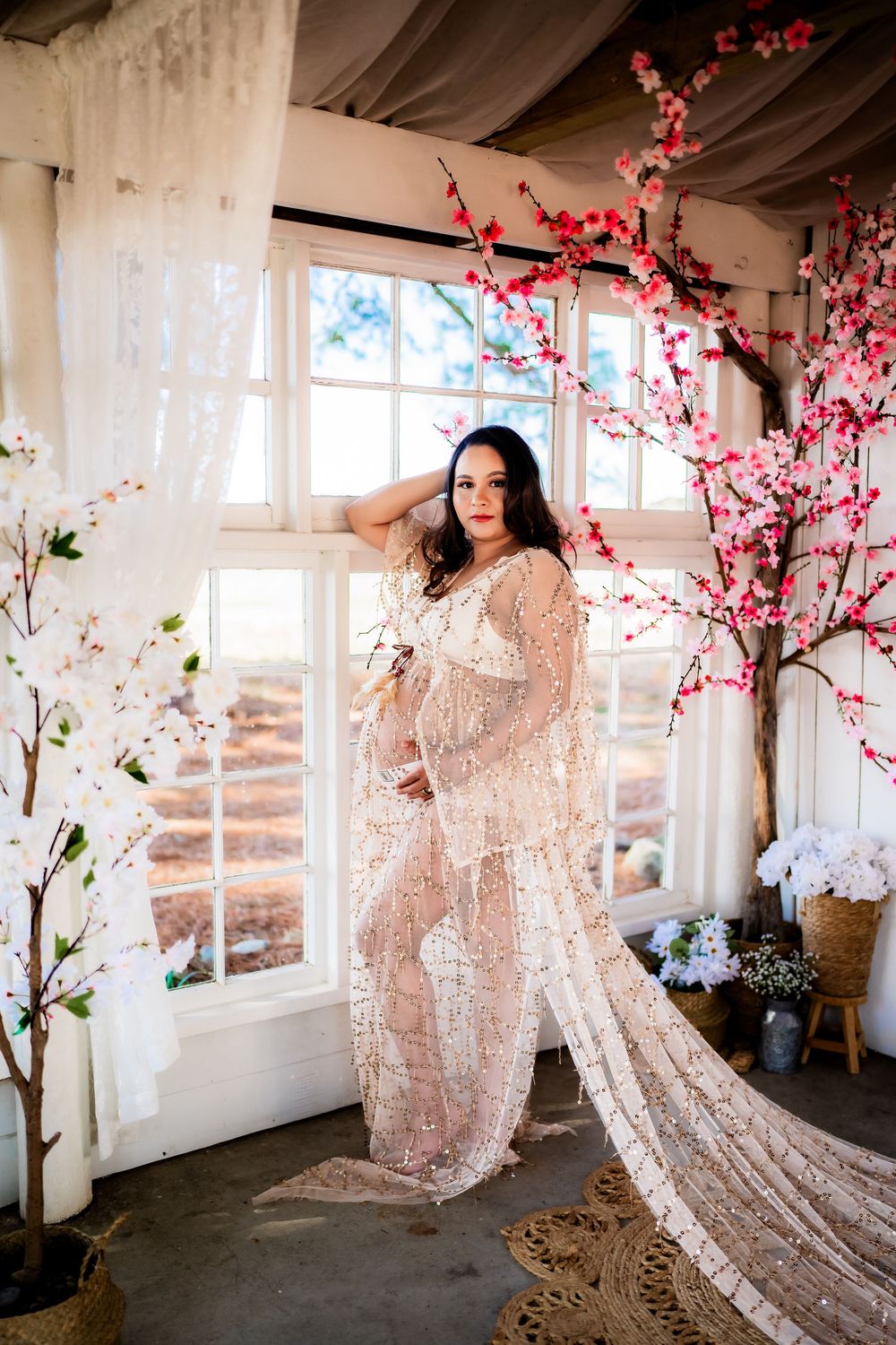 Pregnant woman in lace gown poses by window with cherry blossom trees in elegant studio maternity photo.