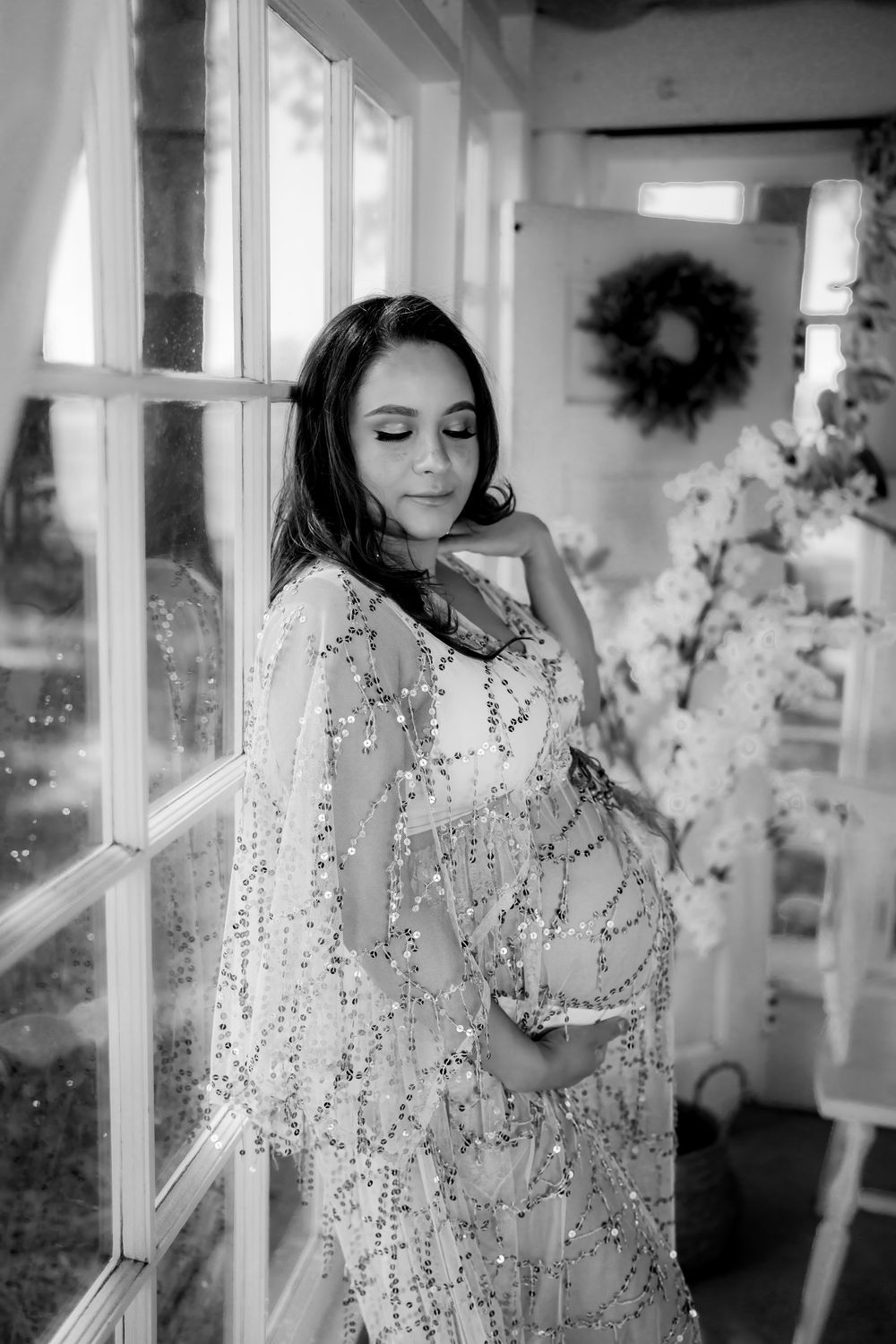 Black and white maternity photo of a pregnant woman in a sequined gown posing by a window with soft natural light.