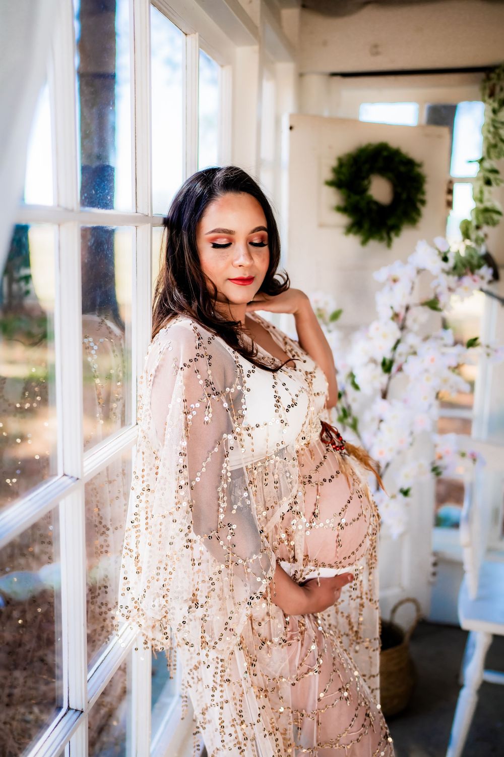 Pregnant woman in gold sequin gown poses by window with floral decor.