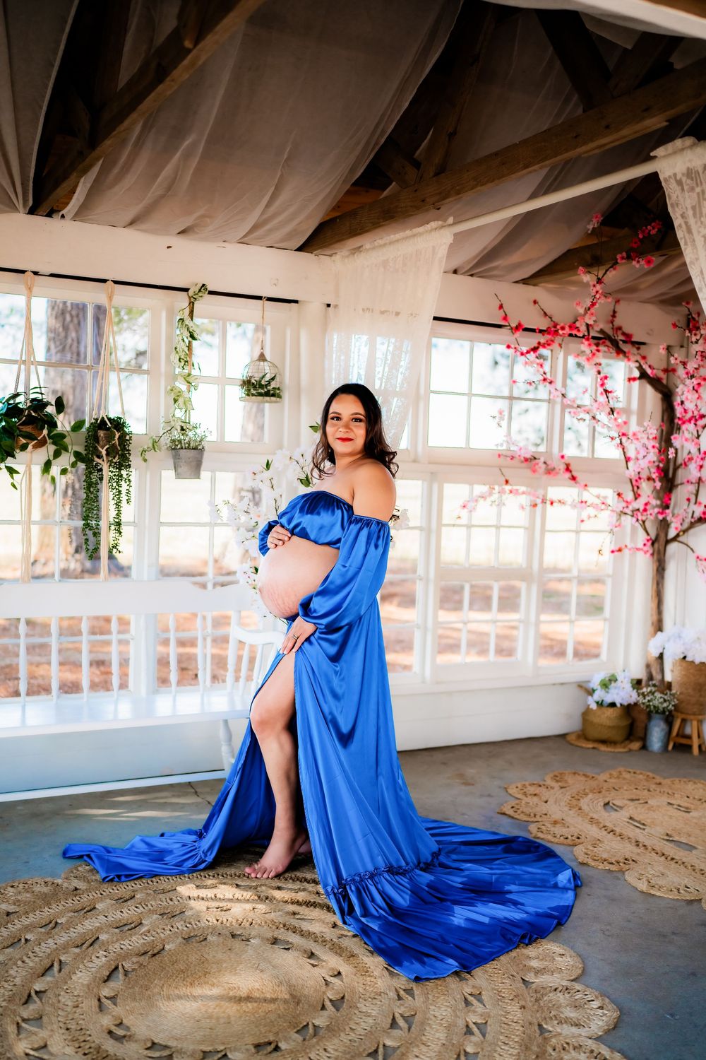 Pregnant woman in elegant blue off-shoulder gown with train poses in bright studio with cherry blossom decor.