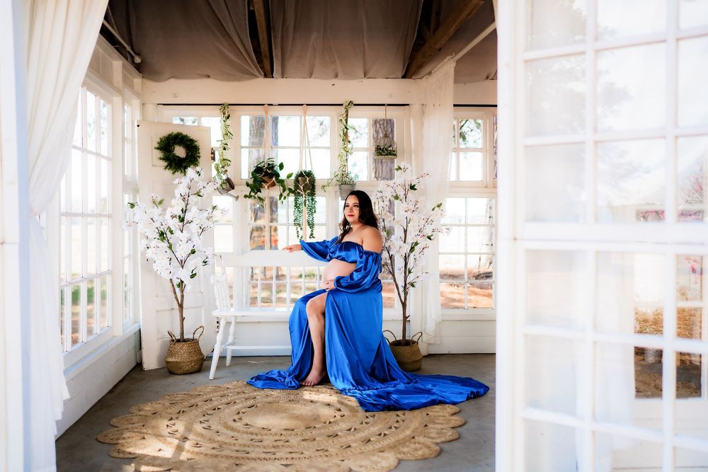 Pregnant woman in royal blue off-shoulder gown seated in elegant white sunroom with floral decor and natural light.