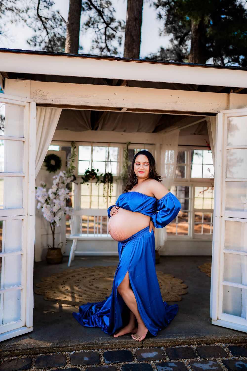Pregnant woman in blue satin off-shoulder gown cradling baby bump, standing in white French door entrance.