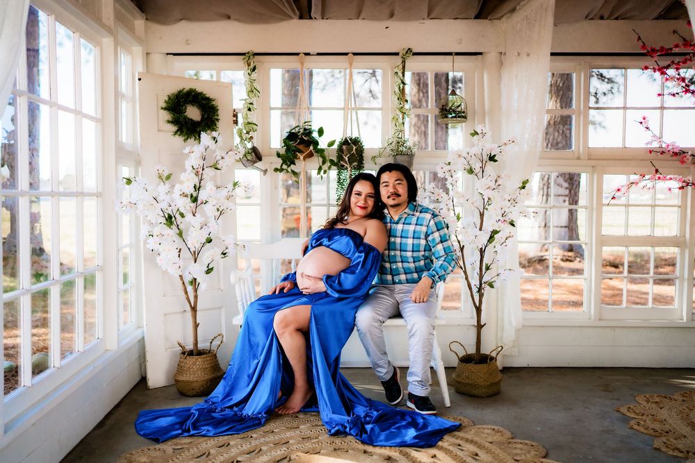 Pregnant woman in blue gown with partner in sunlit studio decorated with white trees and floral wreaths for maternity shoot.