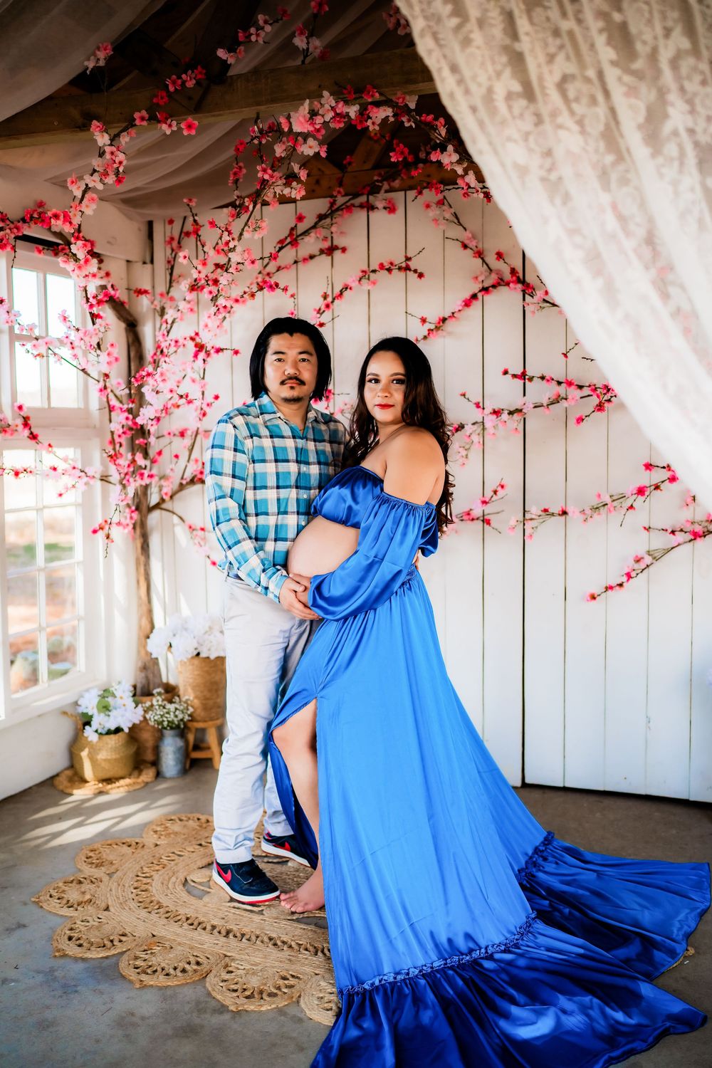 Couple poses for maternity photo shoot in blue gown with cherry blossom backdrop.