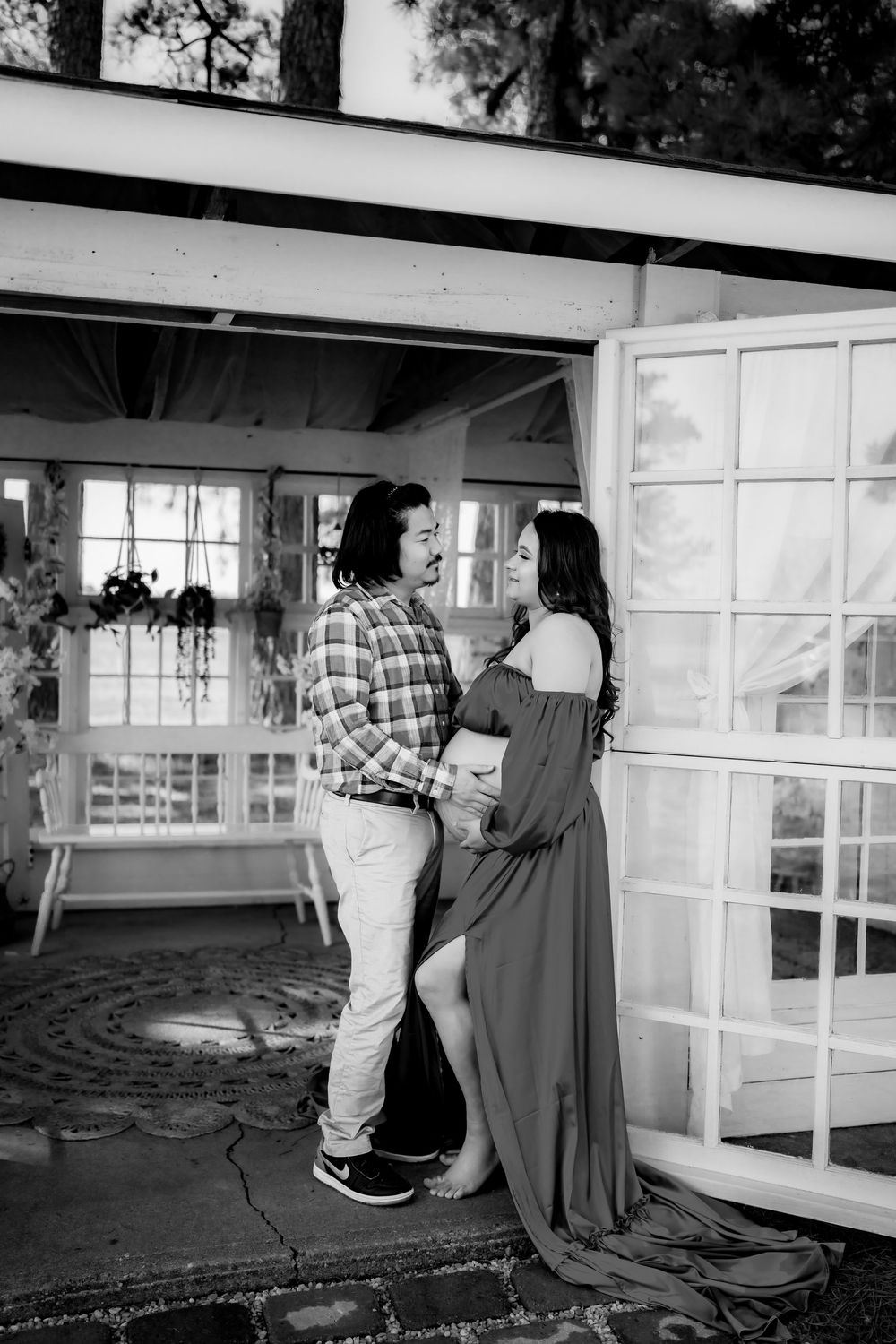 Black and white photo of a couple embracing near white French doors during a maternity photoshoot.