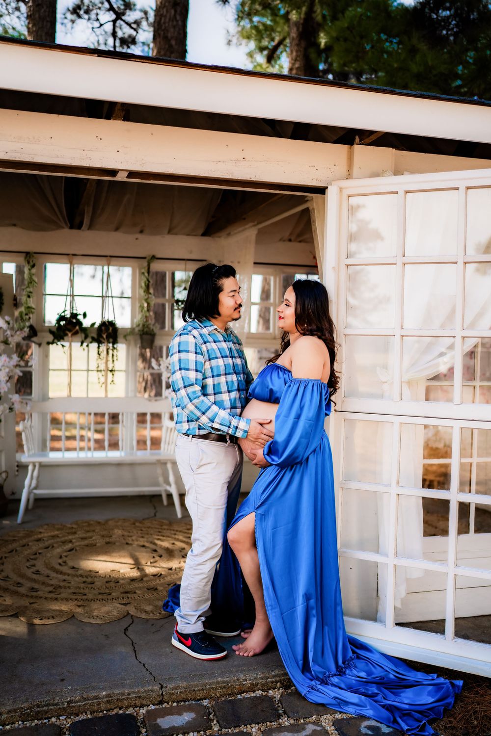 Pregnant woman in blue off-shoulder gown poses with partner near white French doors outdoors.