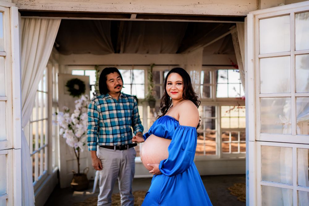 Couple poses for maternity photo shoot, woman in blue off-shoulder gown showing baby bump, man in plaid shirt holding her hand.