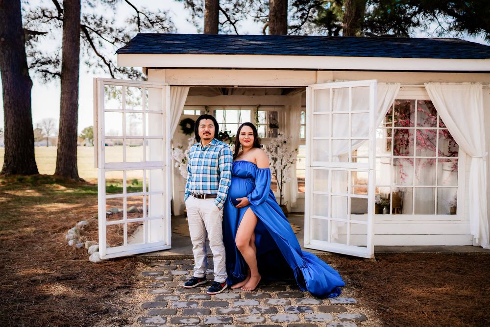 Couple poses in front of white cottage doors, woman in blue maternity gown, man in plaid shirt, outdoor maternity photo.