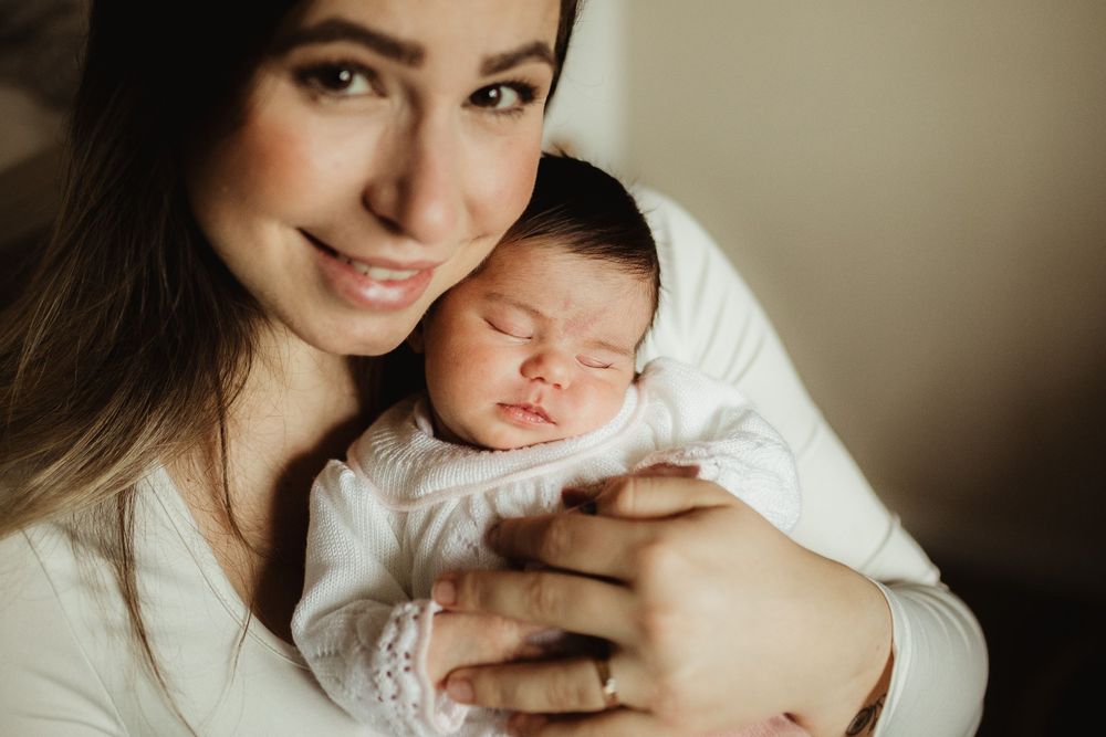 A smiling mother holds her sleeping newborn baby dressed in white against a soft, neutral background.