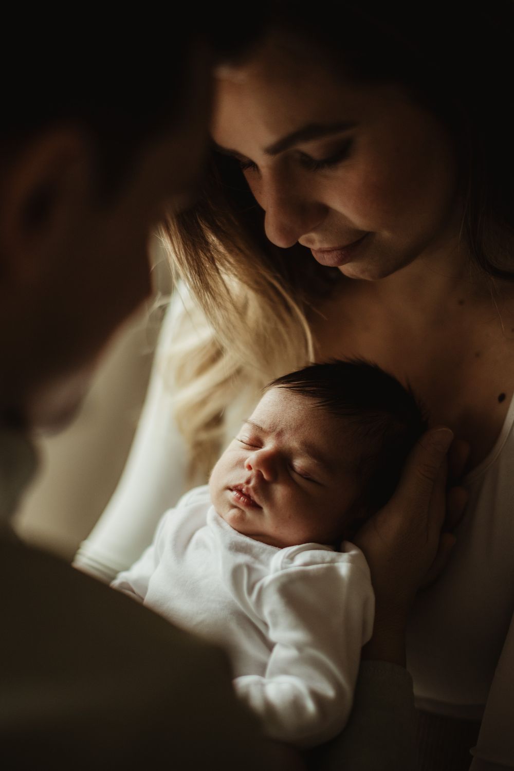 Parents gazing lovingly at their sleeping newborn baby in a tender, dimly lit portrait.