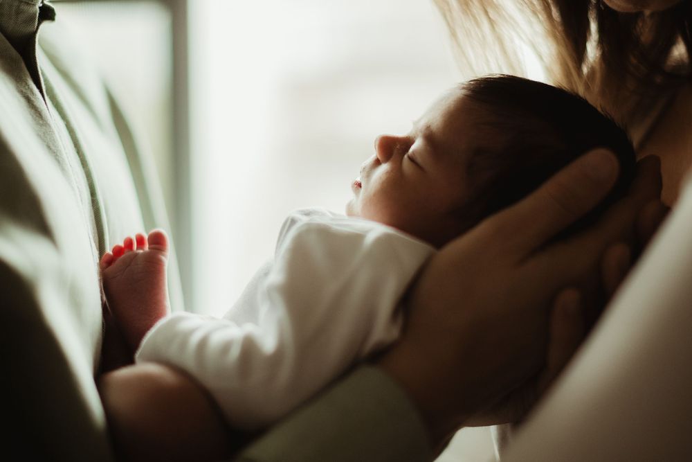 Newborn baby sleeping peacefully in parent's arms near a bright window in soft light.