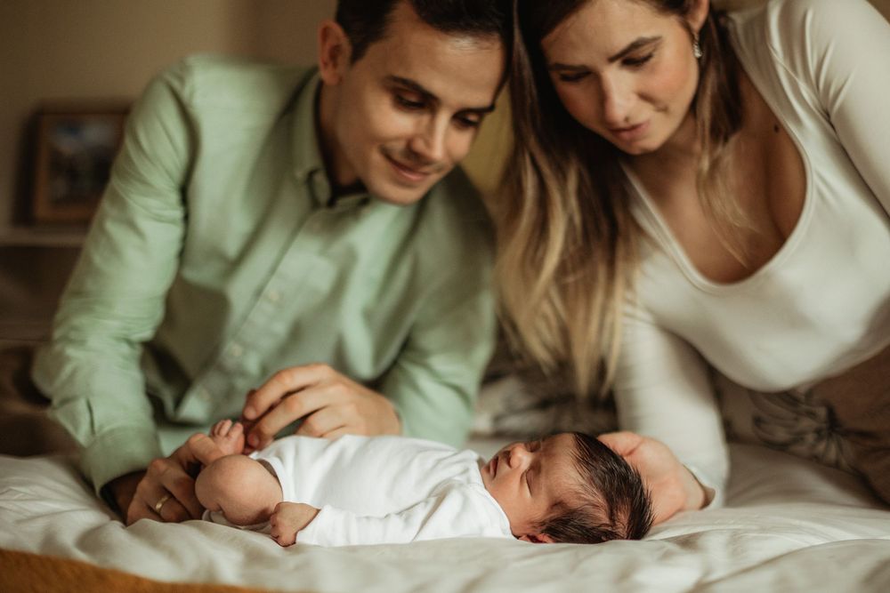 Young parents smiling and bonding with their swaddled newborn baby lying on a bed.