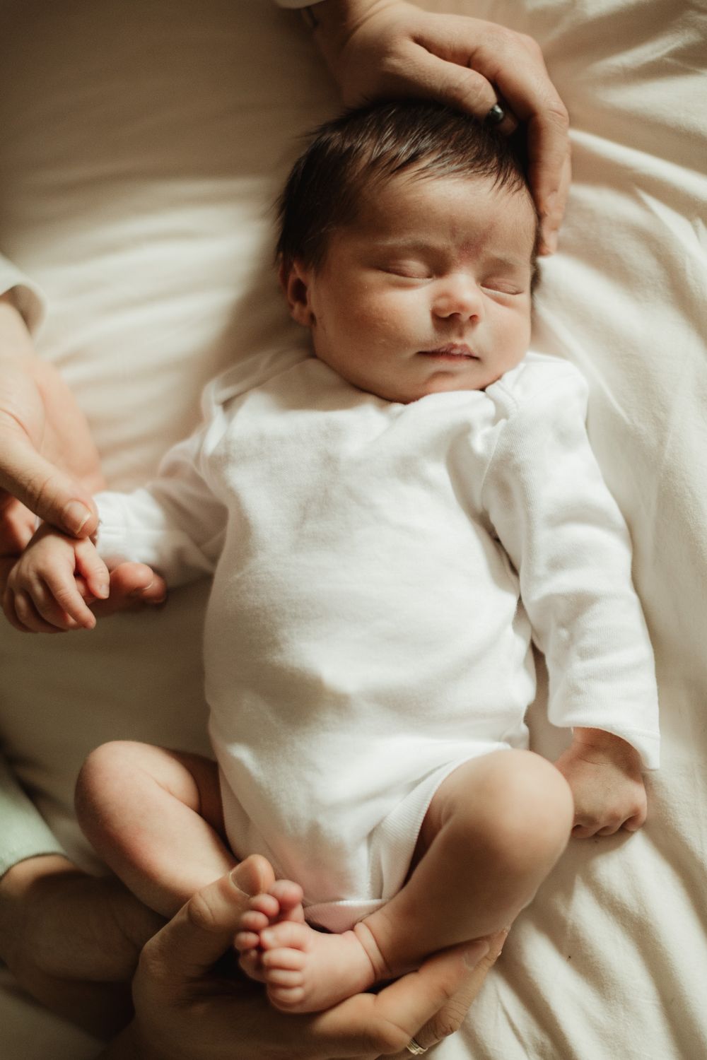 Newborn baby sleeping peacefully in white onesie while parent gently cradles head on soft white bedding.