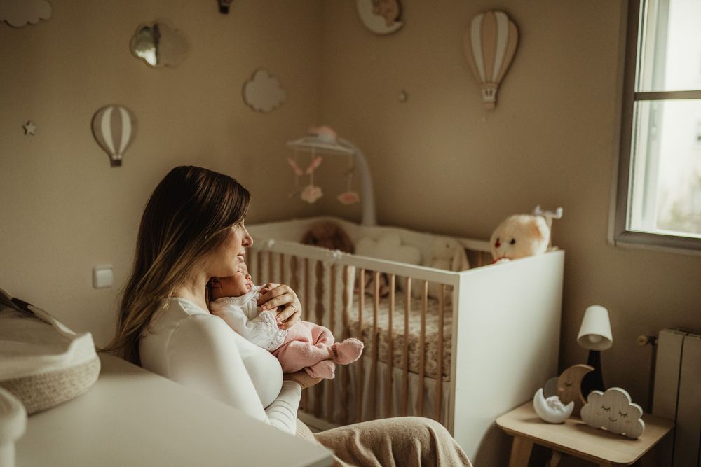 Mother holding newborn baby in a softly decorated nursery with white crib, pastel decor, and hot air balloon wall ornaments.