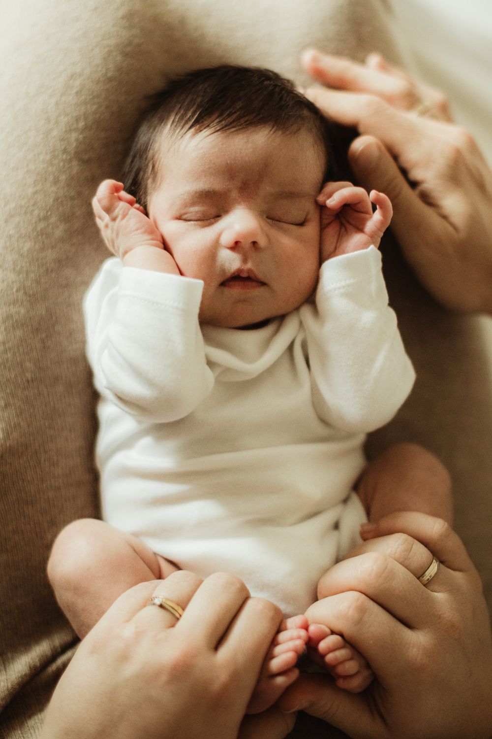 Sleeping newborn baby in white onesie cradled in parents hands, peaceful infant with dark hair.
