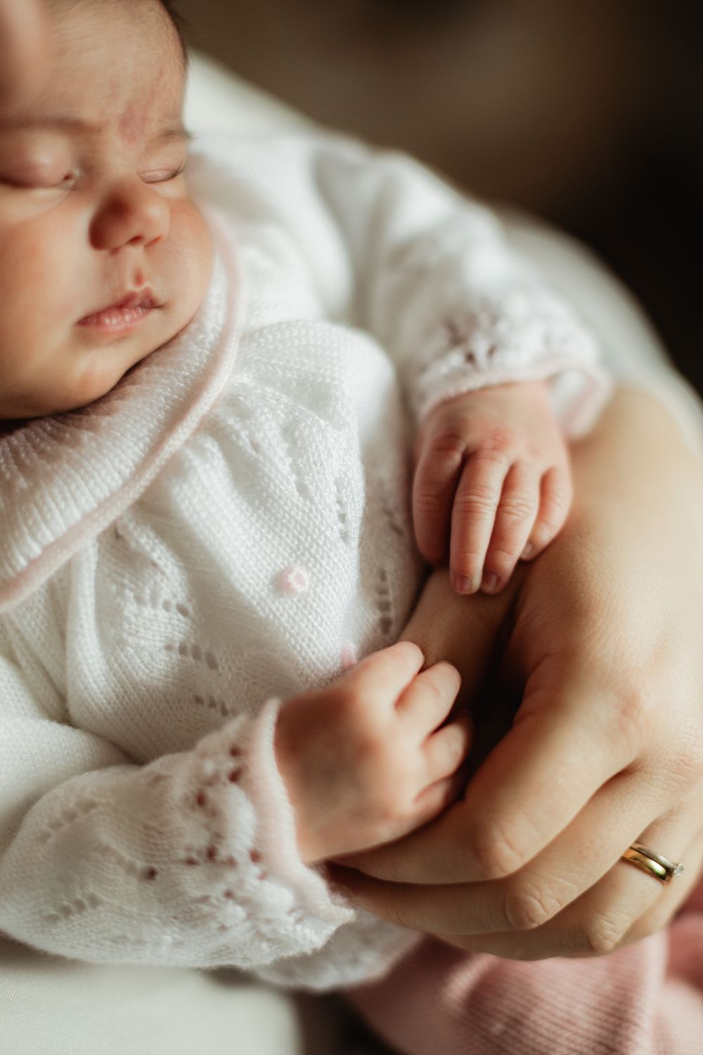 Sleeping newborn baby in white knit outfit held gently by parent wearing gold wedding ring.