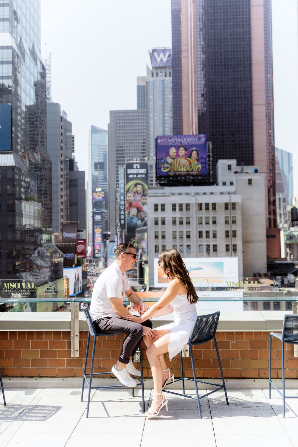 A couple sits at an outdoor rooftop table with city buildings and skyscrapers in the background.