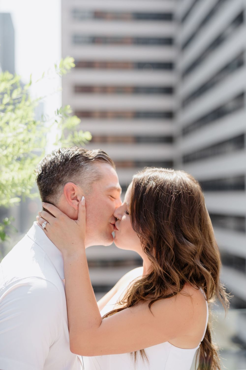 A romantic couple embraces in an urban setting with modern buildings in the background.