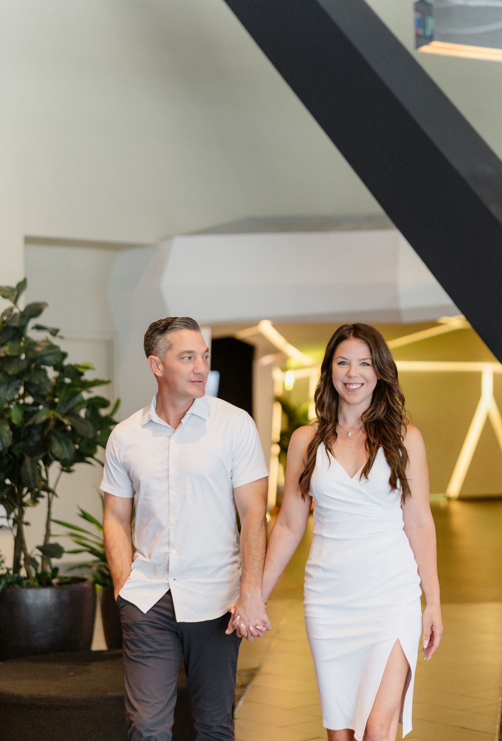 A couple in matching white outfits walks playfully through a modern indoor space with plants.