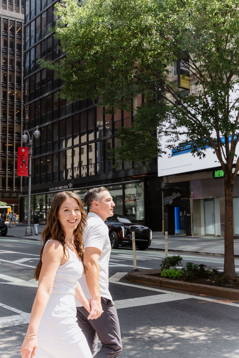 A couple in white clothing strolls together on a sunny city street lined with buildings and trees.