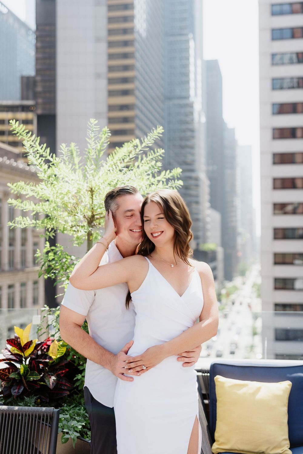 Couple in white outfits embracing on a rooftop terrace with urban skyline and greenery.