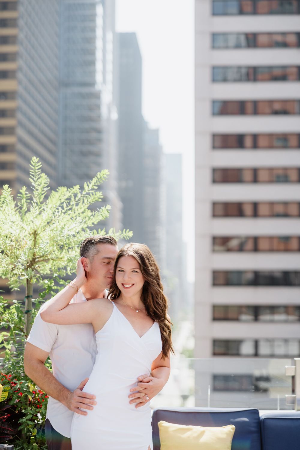 A couple in white outfits pose together on a rooftop terrace with urban skyscrapers in the background.