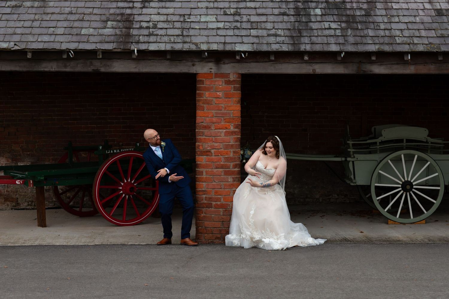 Wedding couple poses near brick building with vintage wagon wheels.