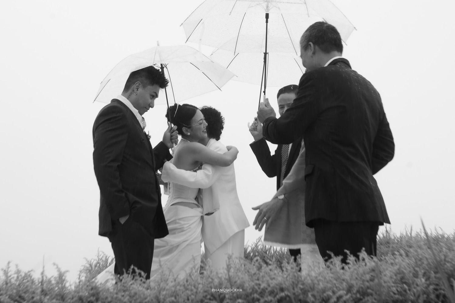 Wedding party shares emotional embrace under clear umbrellas in misty field, black and white photo.