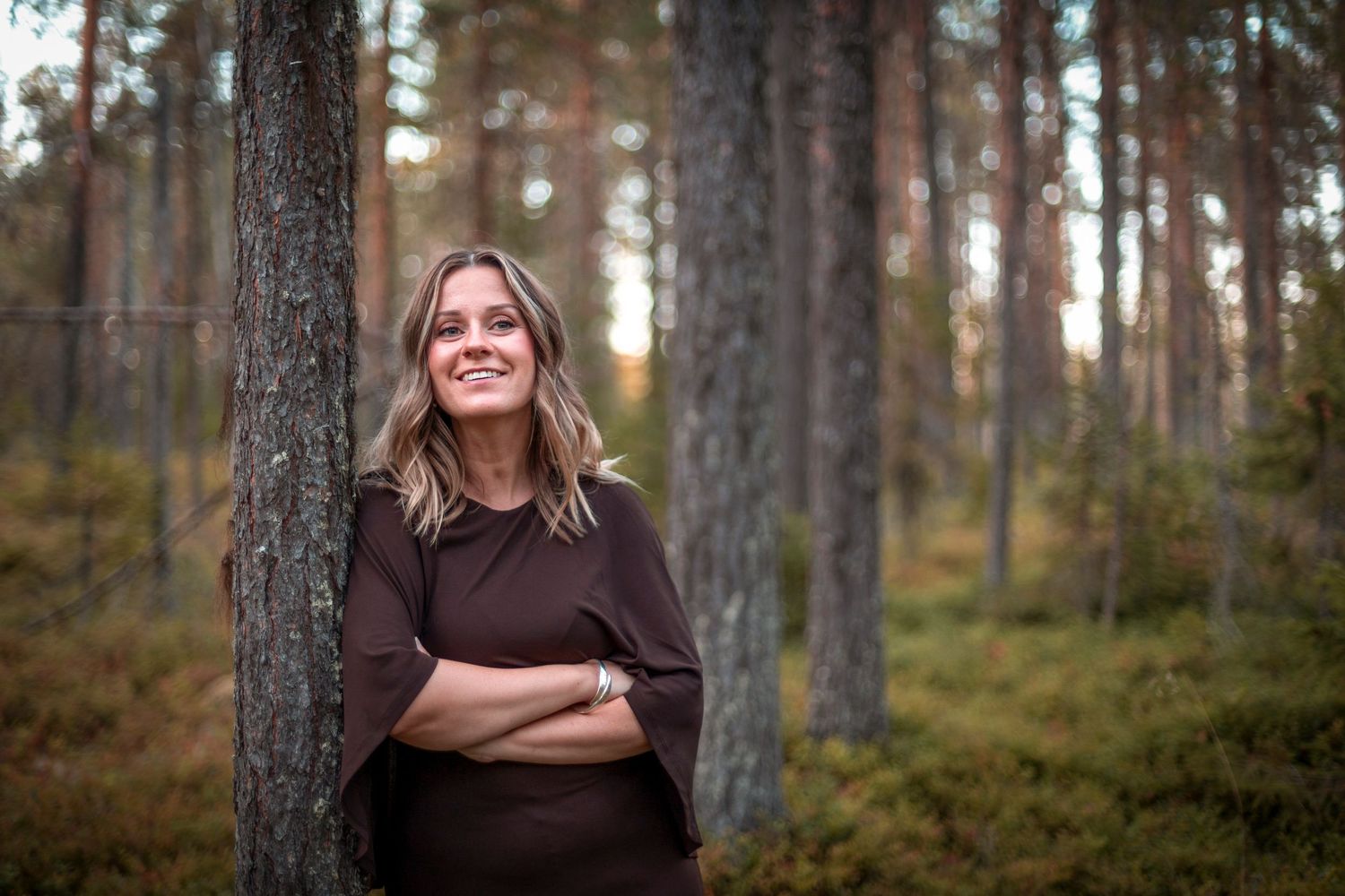 Smiling woman with blonde hair leaning against a pine tree in a sunlit Scandinavian forest.