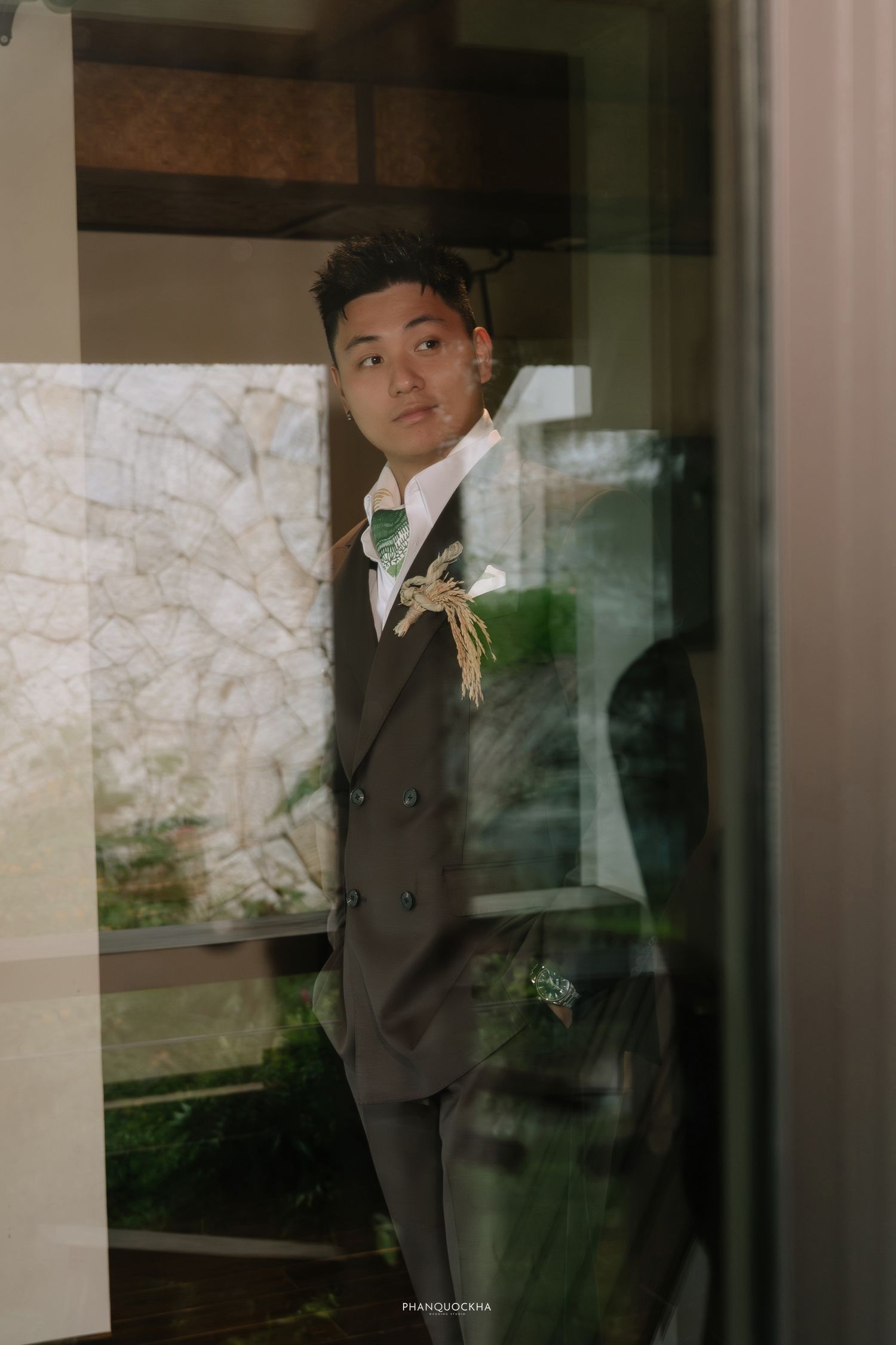 Groom in dark suit with green boutonniere reflected in glass door at wedding venue.