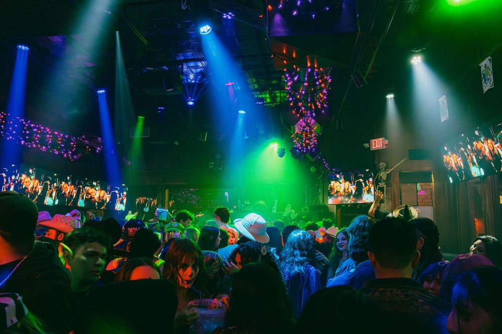 Vibrant blue and green stage lights illuminate a dark nightclub venue with dancing silhouettes and colorful decorations.