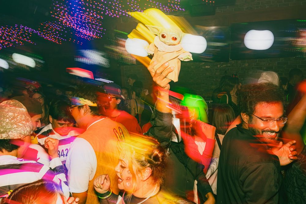 Blurred motion of people dancing and moving in a dimly lit nightclub with colorful lights and DJ equipment.