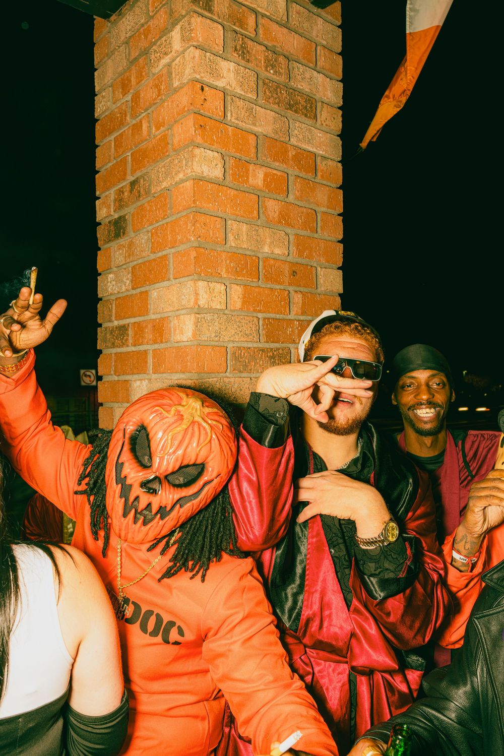 Group of people in Halloween costumes including someone in bright orange jumpsuit with scary mask posing at night.