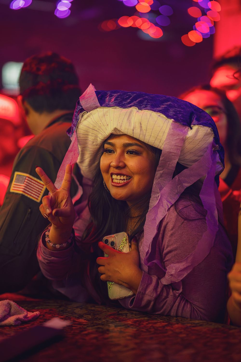 A person wearing a purple costume with white headpiece makes a peace sign at a nightclub with red lighting.
