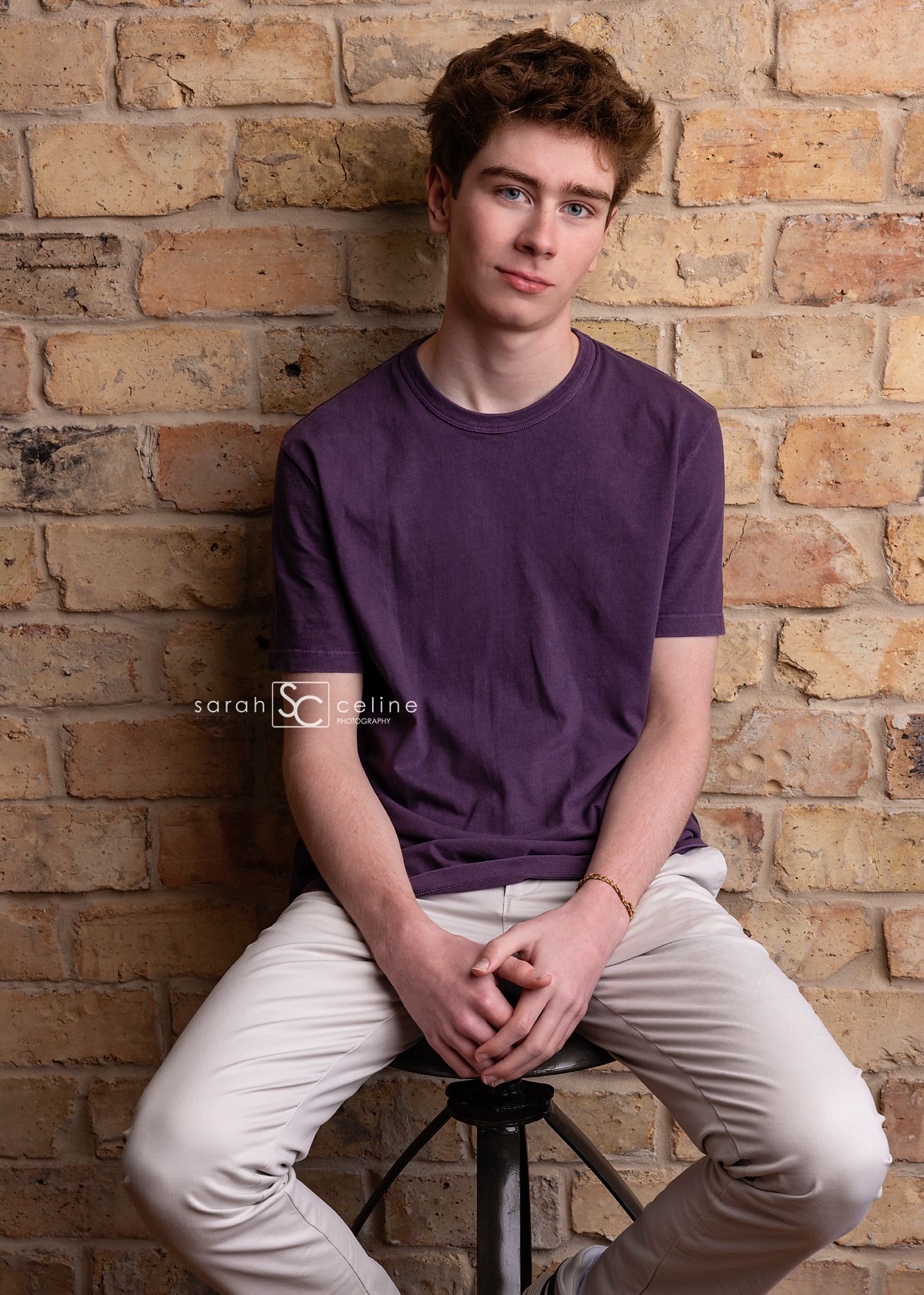 A casual portrait taken against a rustic brick wall background with the subject wearing a purple t-shirt and khaki pants.