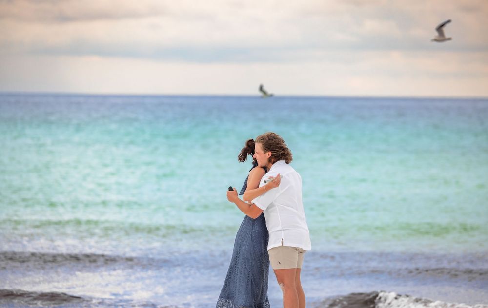 A romantic couple embraces on a beach with turquoise waters and cloudy skies in the background.
