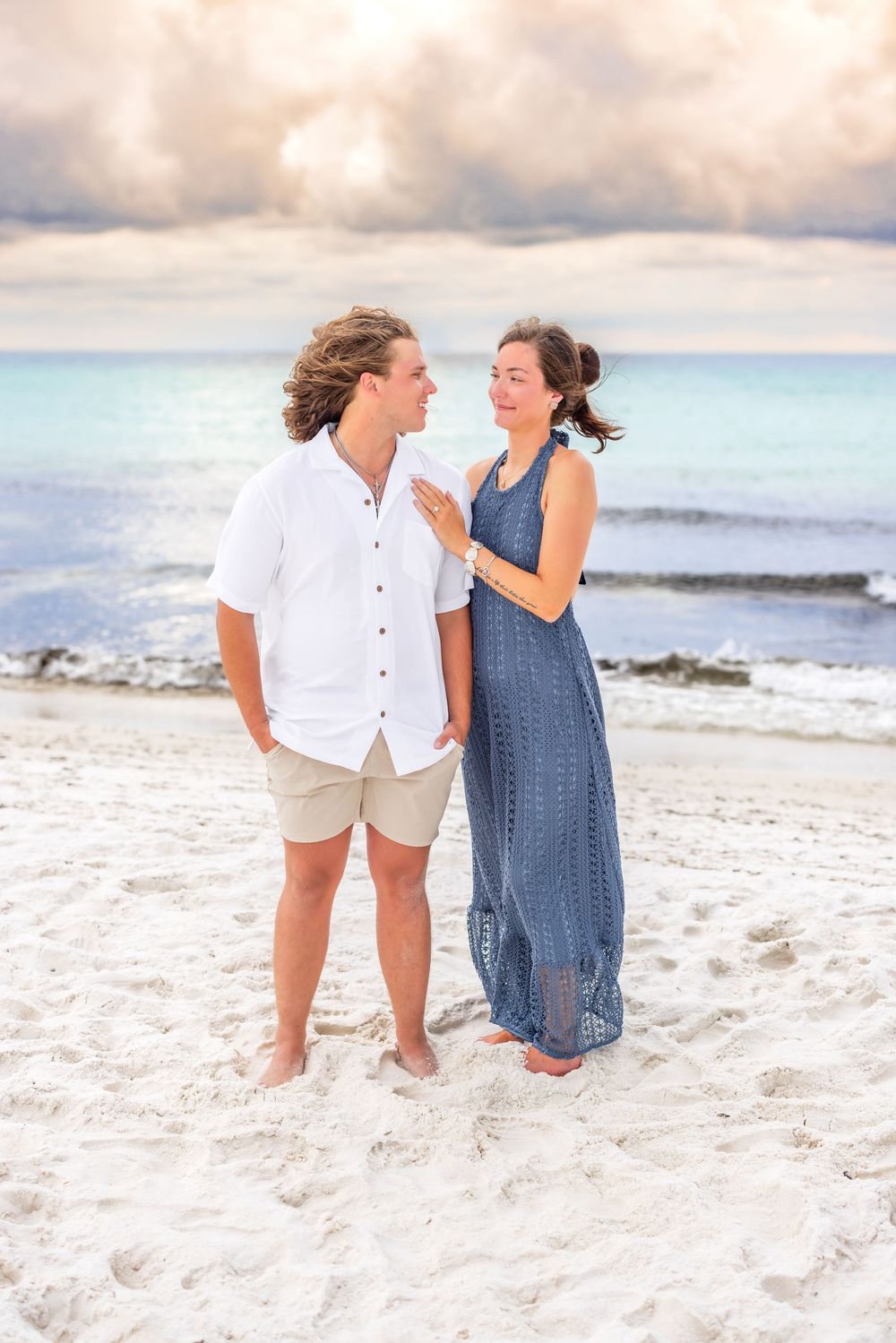 Two people walking together on a white sandy beach with gentle waves and cloudy skies in the background.