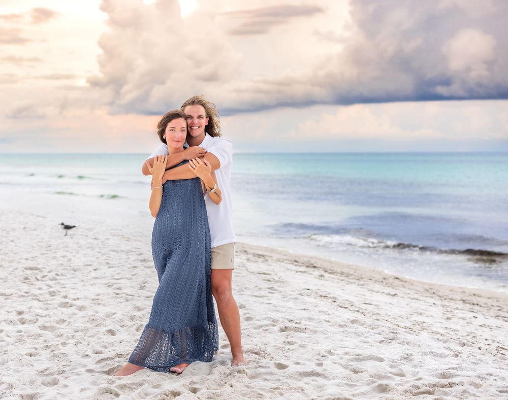 People embrace on a scenic white sandy beach with gentle waves and cloudy skies at sunset.