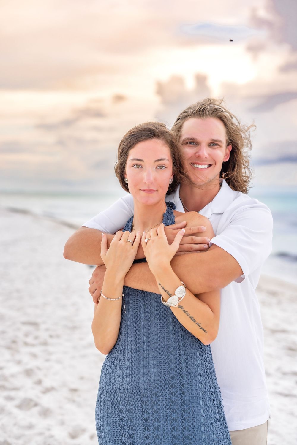 Two people embrace on a white sandy beach during sunset as warm light illuminates the peaceful coastal scene.