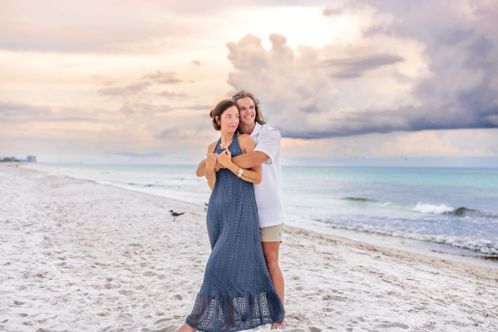 Happy couple embraces on a scenic beach during sunset with ocean waves and cloudy sky in the background.