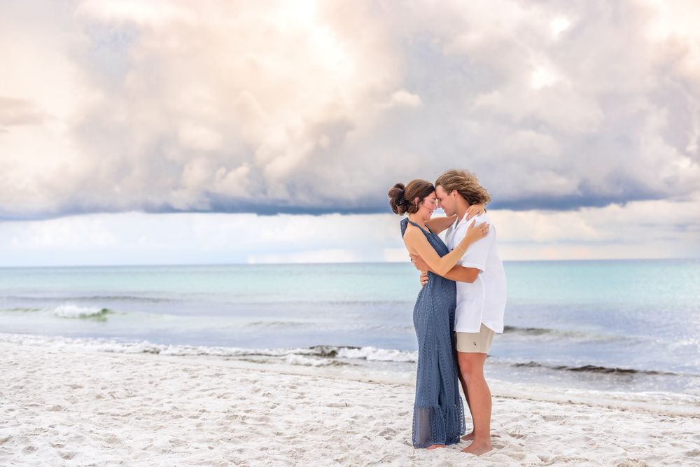A couple embraces on a scenic beach with turquoise waters and dramatic cloudy skies in the background.