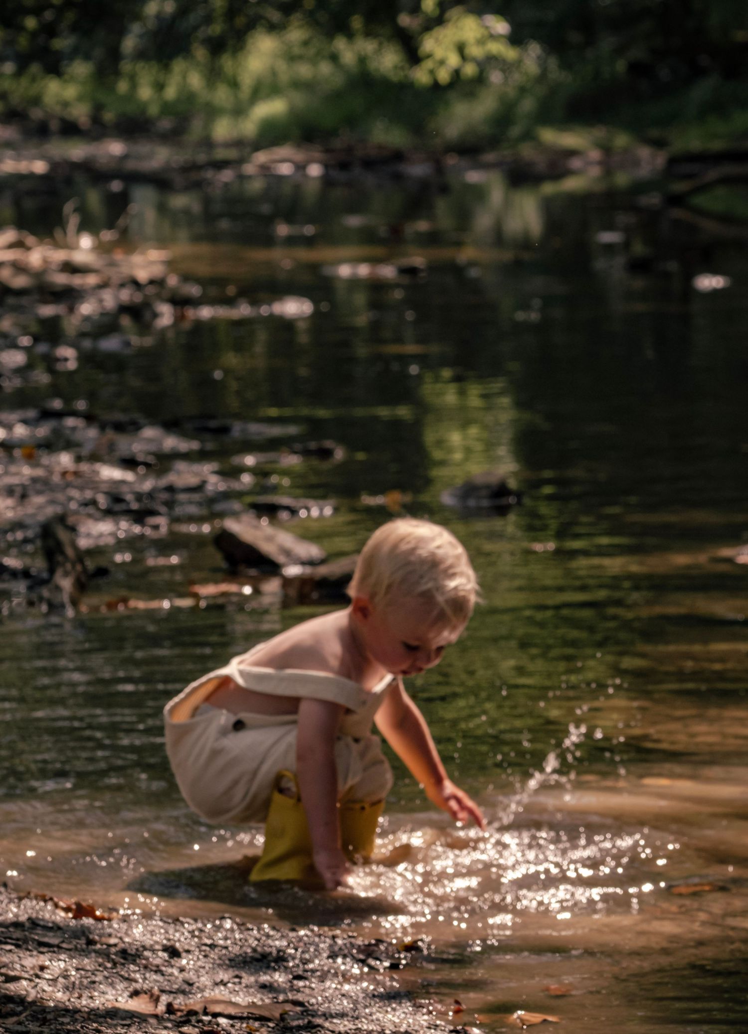 Young child crouches by a river's edge playing in the sparkling water on a sunny summer day.