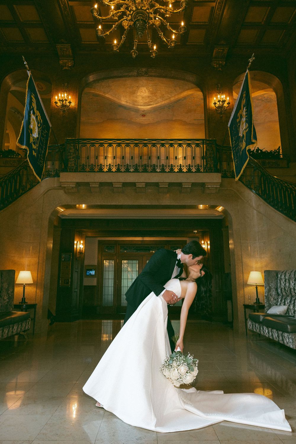 A romantic wedding couple shares a dramatic dip pose in an elegant historic venue with chandeliers and grand staircase.