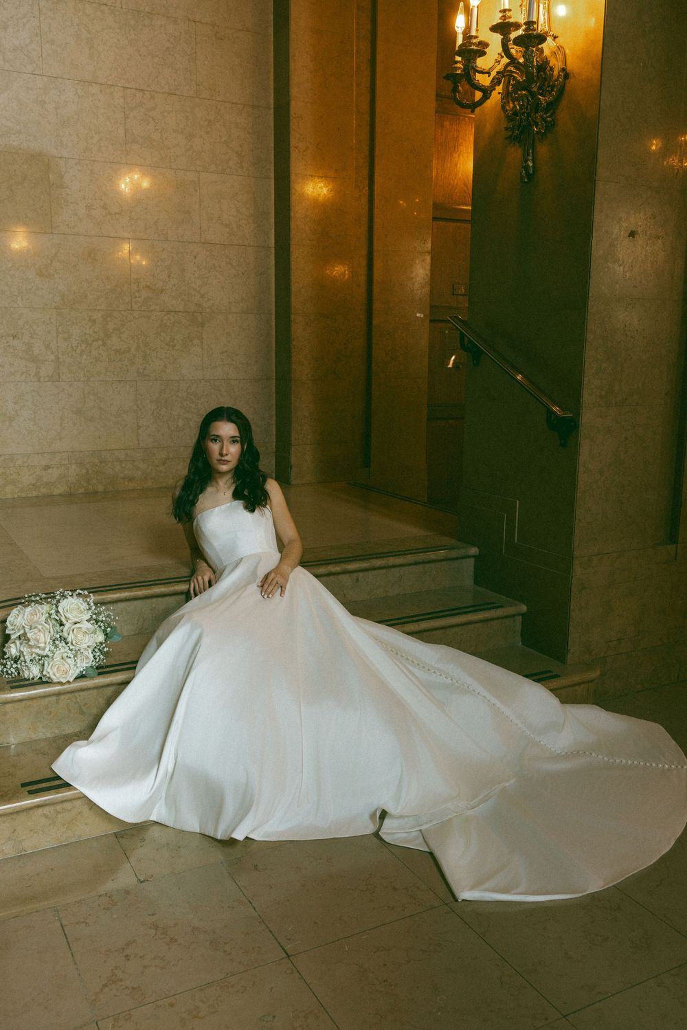 Bride in an elegant white ballgown poses gracefully on marble stairs with a white floral bouquet.