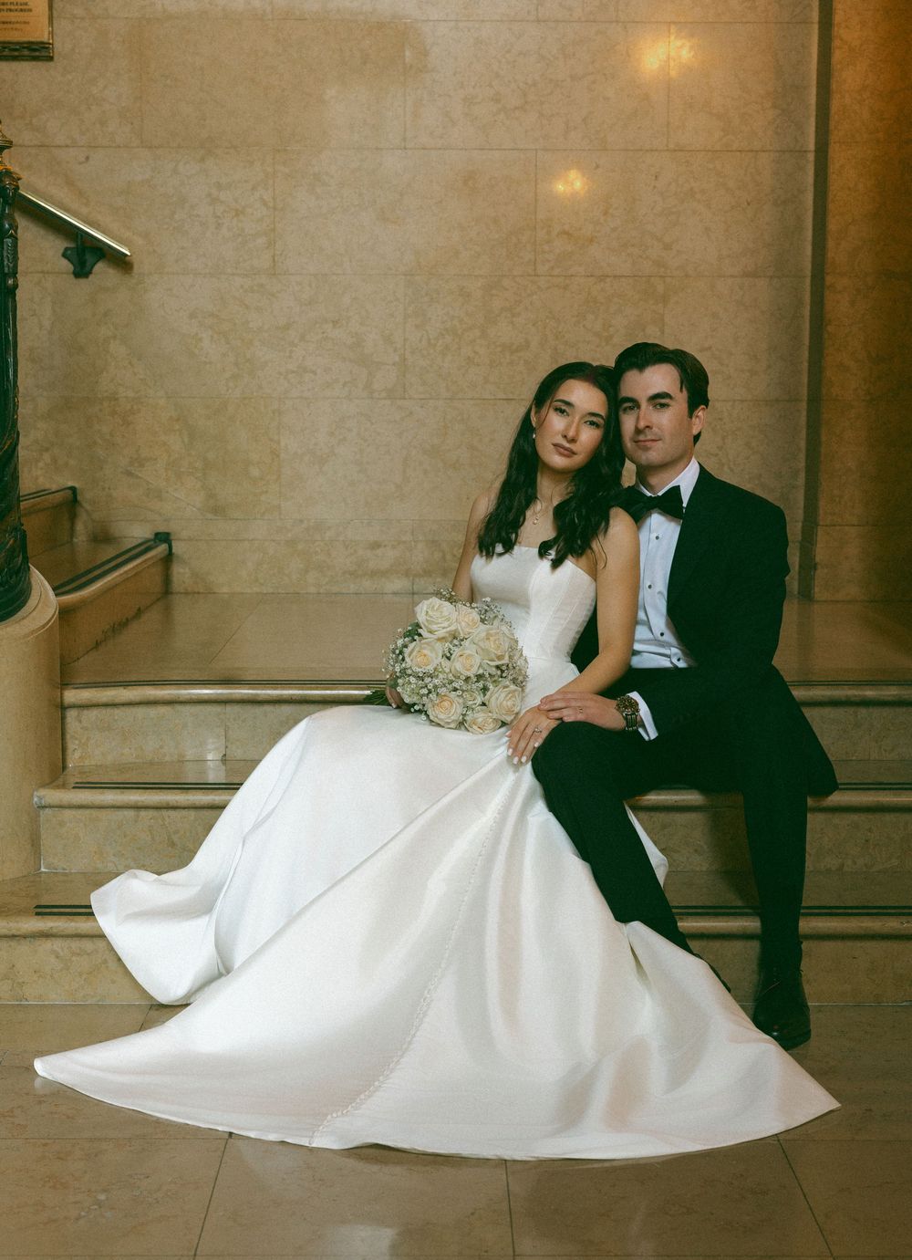 Newlywed couple sits together on stone steps, bride in flowing white gown holding a bouquet.